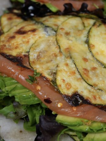 A freshly prepared courgette wrap filled with smoked salmon, avocado slices, cream cheese, and salad leaves, garnished with thyme leaves and chilli flakes.