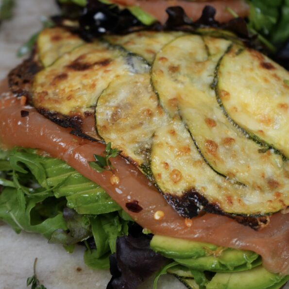 A freshly prepared courgette wrap filled with smoked salmon, avocado slices, cream cheese, and salad leaves, garnished with thyme leaves and chilli flakes.