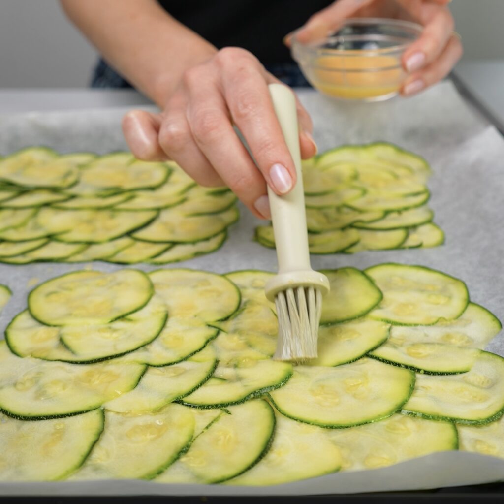 Courgette slices on a baking tray being brushed with egg wash.