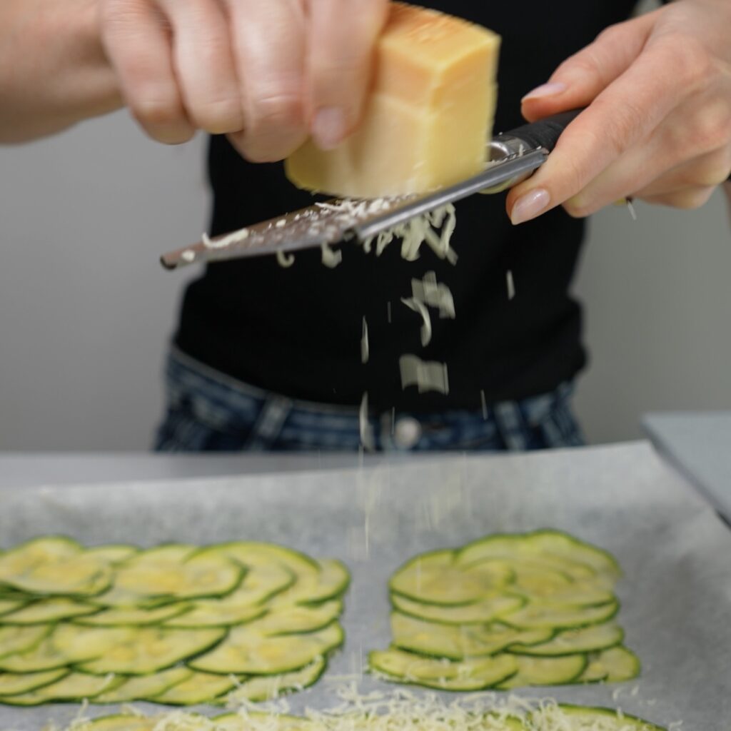 Grated Parmesan cheese being sprinkled over courgette slices on a baking tray.