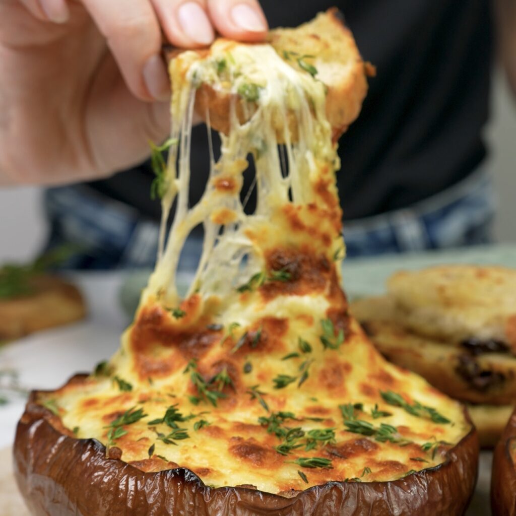 Crostini being dipped into twice-baked aubergine dip topped with melted mozzarella and fresh herbs