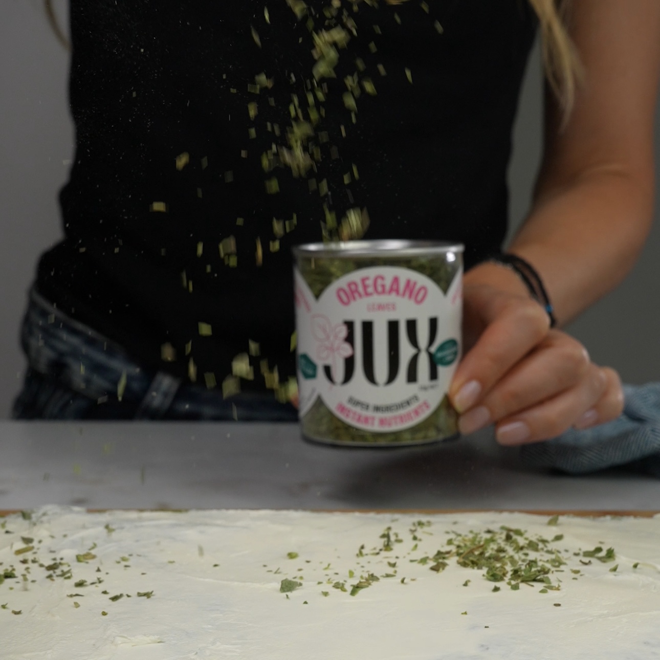 Oregano being sprinkled over cream cheese spread on a spinach roll base.