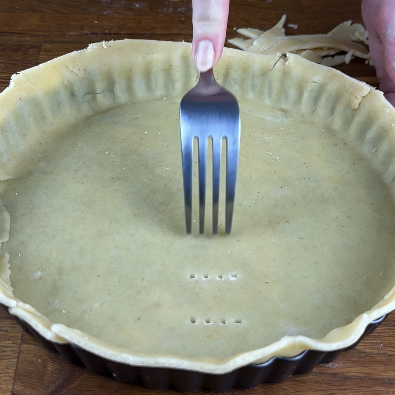 A photo showing the bottom of a tart tin lined with raw pastry dough being pricked with a fork to prevent bubbling.