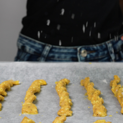 Flaky salt being sprinkled over uncooked Peppery Cheddar Breadsticks on a baking tray.