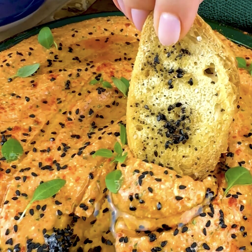 A slice of crusty bread is dipped into a bowl of roasted red pepper and feta dip.