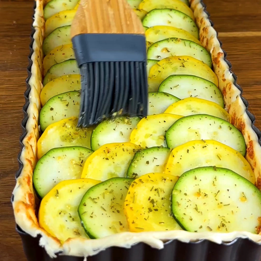 Courgette slices being brushed with oil on a tart.