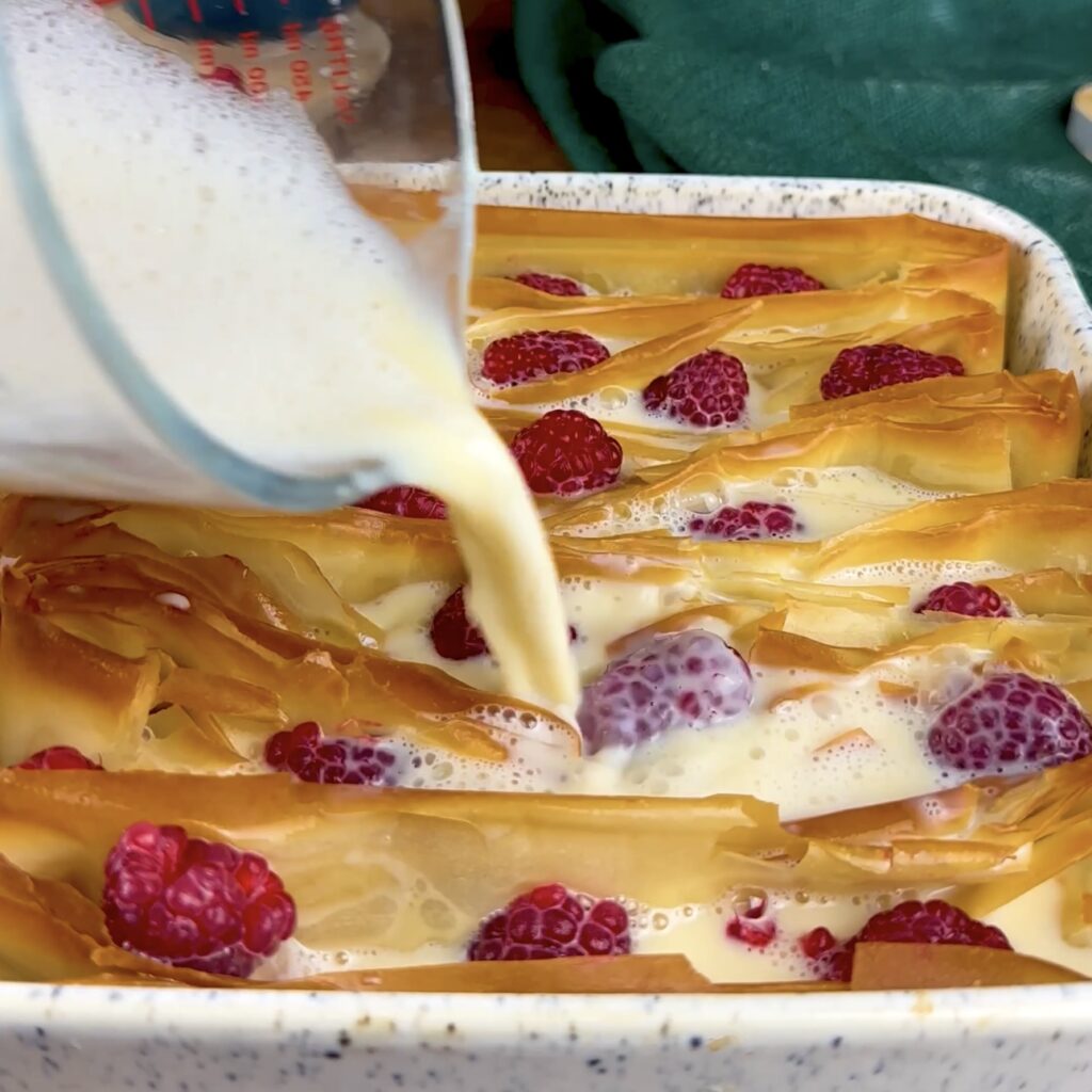 Custard being poured over baked filo pastry with scattered raspberries.