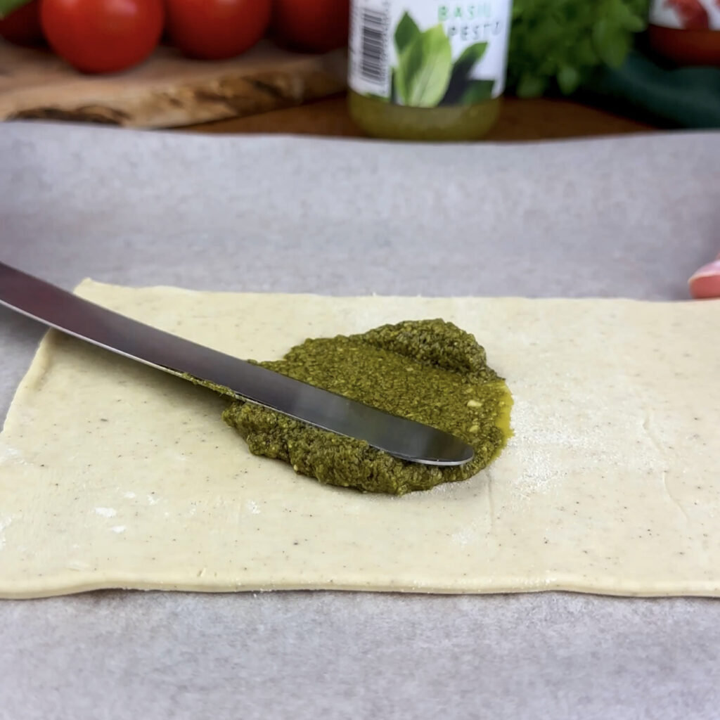 Spreading basil pesto evenly on a section of puff pastry.