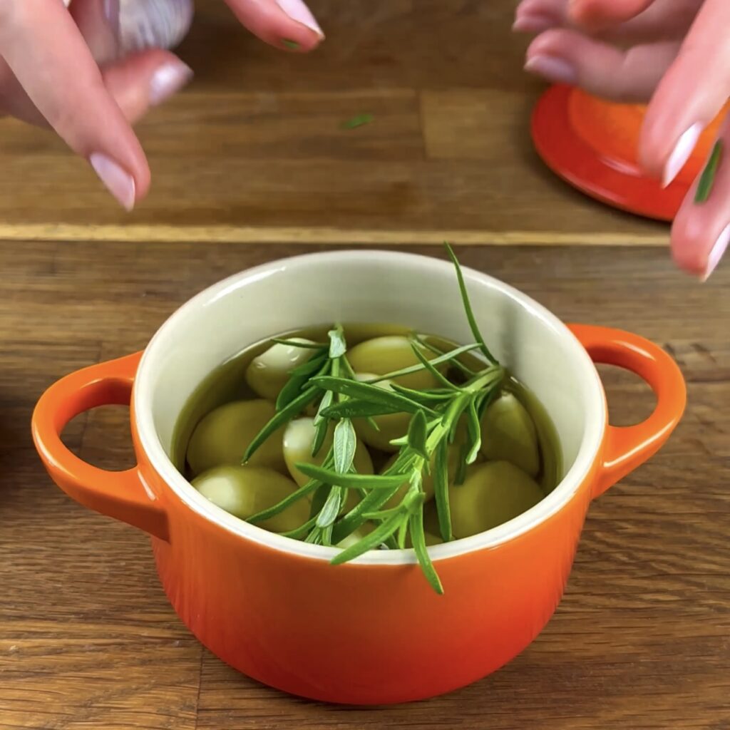 Garlic cloves covered in olive oil and topped with fresh rosemary sprigs, ready for roasting.