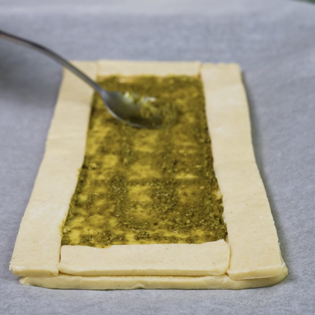 Spreading green pesto evenly over the base of puff pastry rectangles.