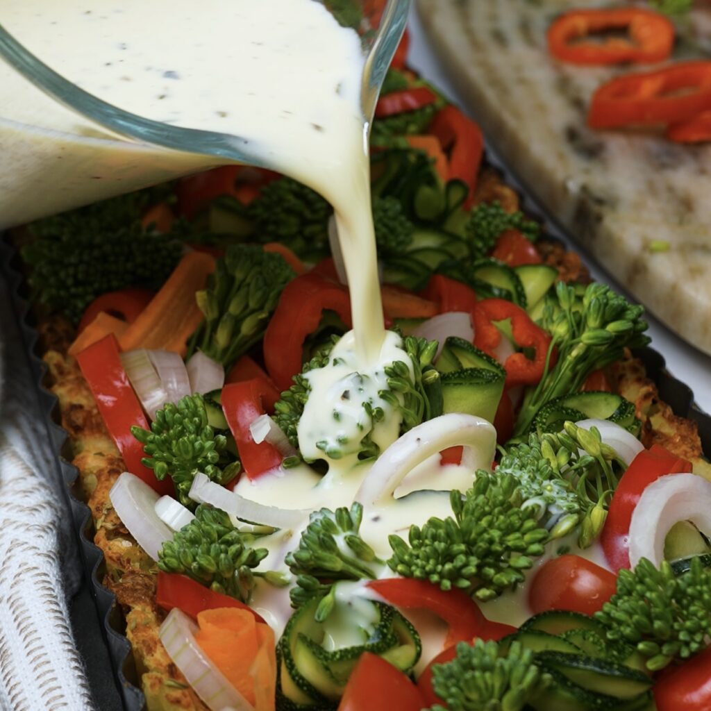 Pouring creamy custard over arranged vegetables in a baked potato crust, ready for baking.