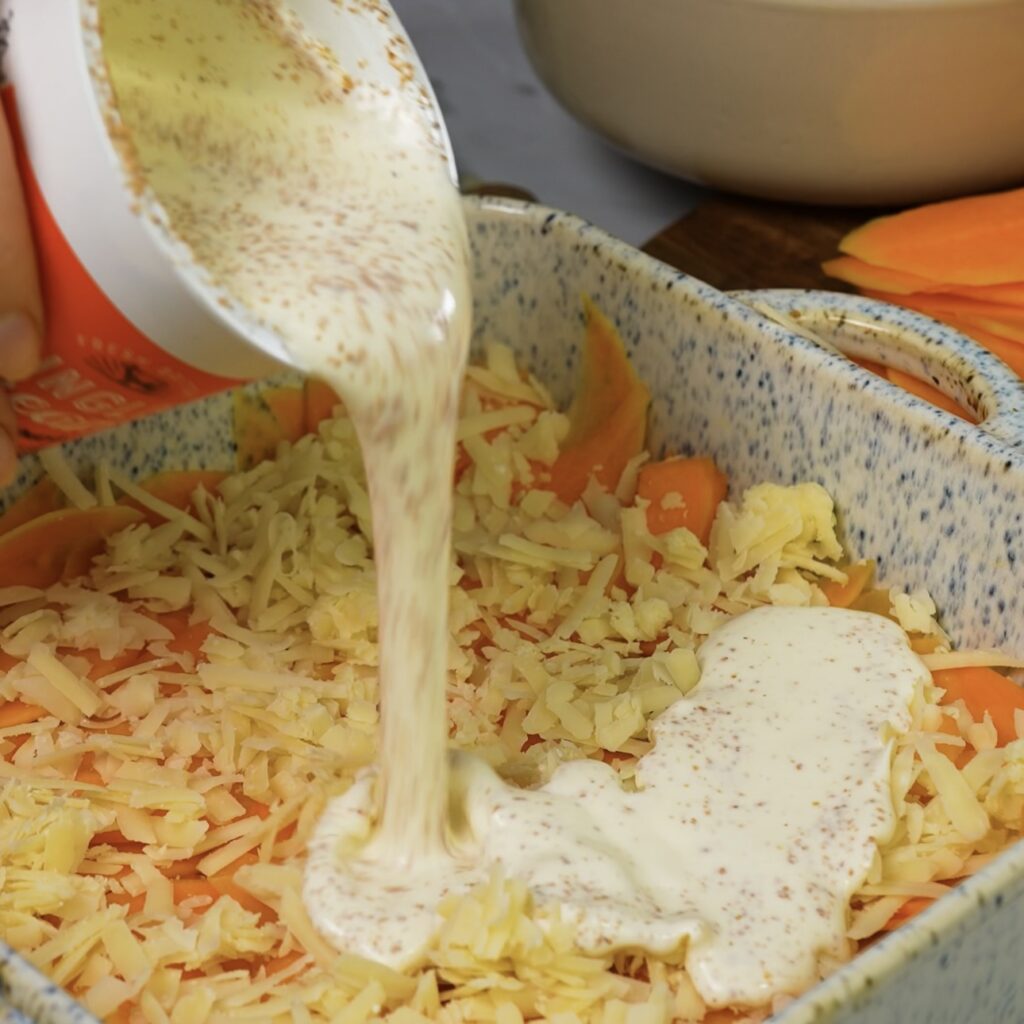 Pouring a creamy mixture over the first layer of butternut squash in a baking dish.