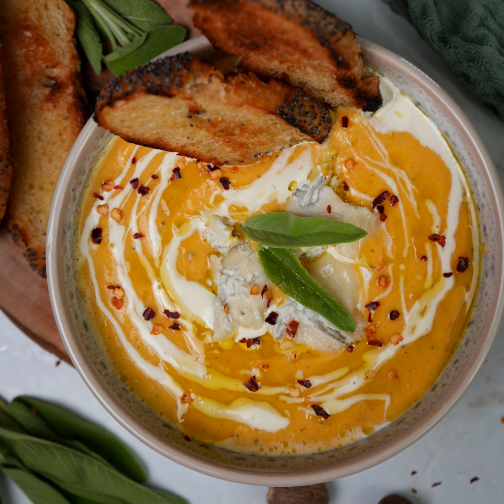 A bowl of Roasted Butternut Squash and Pear Soup topped with crumbled Gorgonzola cheese and a drizzle of olive oil, served with crusty bread on the side.