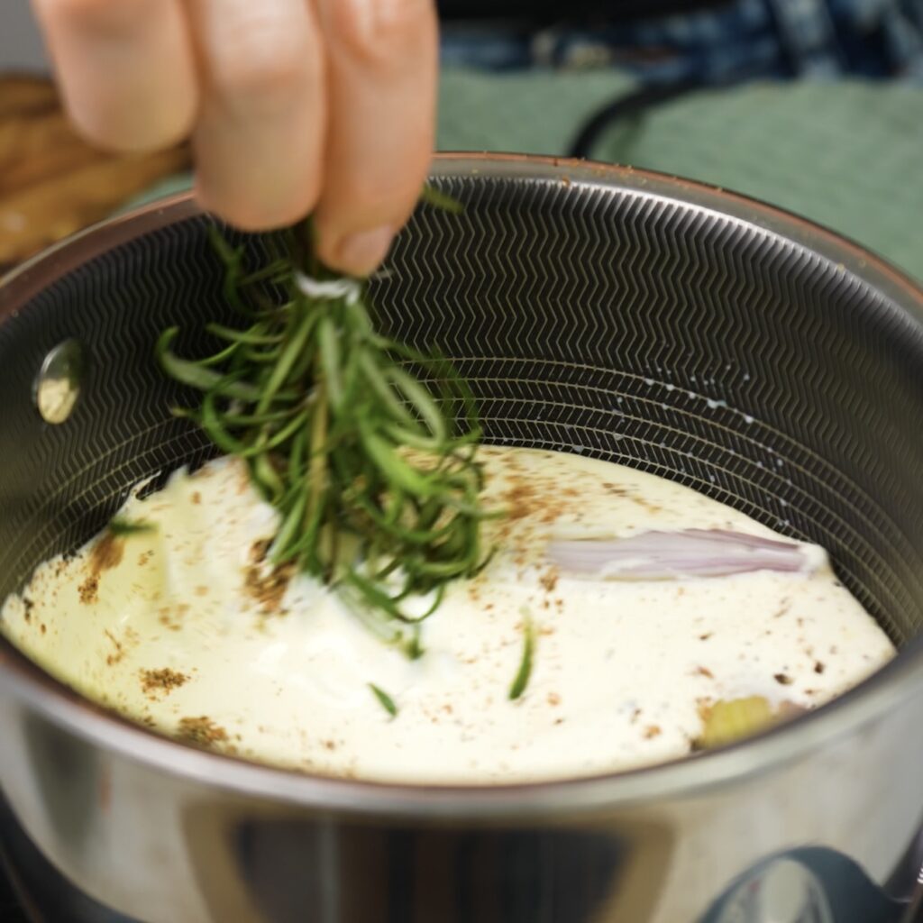 Milk and double cream infusing with rosemary sprigs, halved shallot, and grated nutmeg in a saucepan.