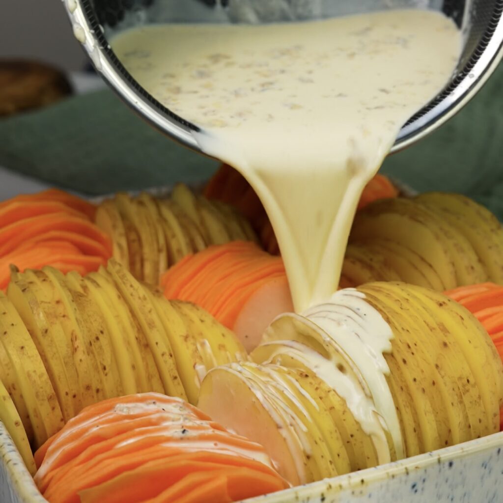 Pouring the infused creamy mixture over layers of sliced sweet and regular potatoes in a baking dish.