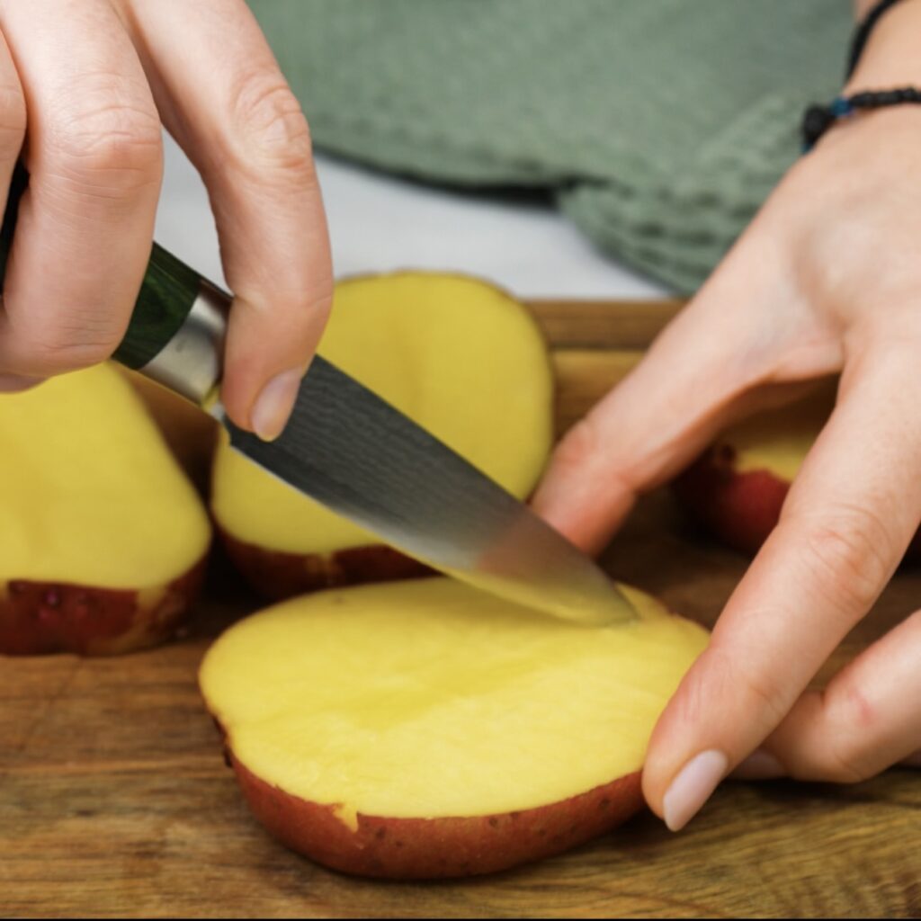 Scoring a potato slice with a knife on a wooden cutting board.