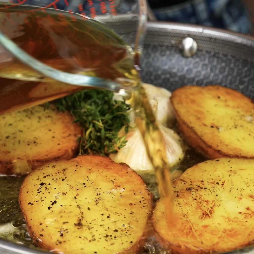 Pouring vegetable stock into a frying pan with golden-brown potato slices.