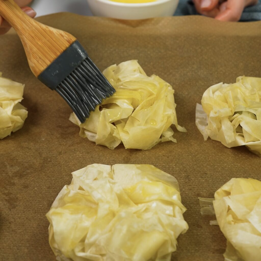 Brushing melted butter over assembled filo buns before baking.