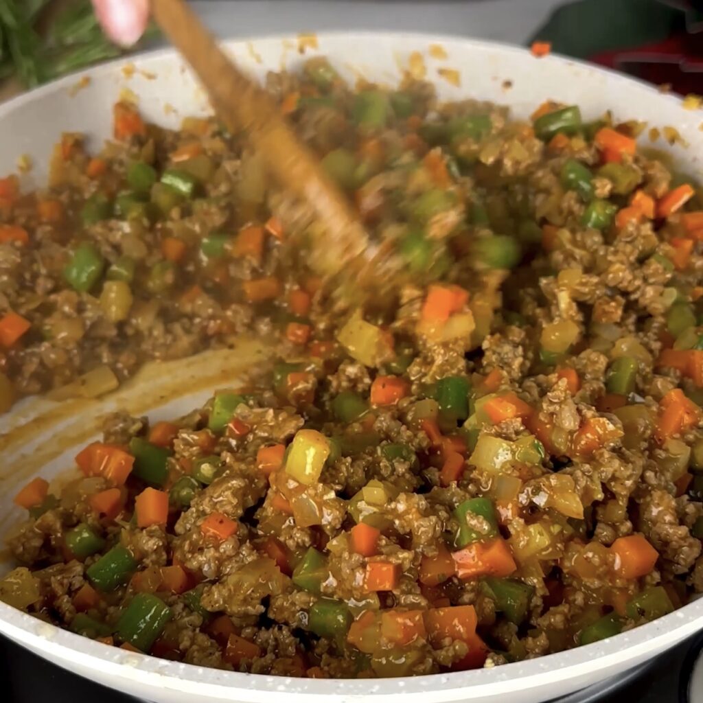 Minced beef and vegetables simmering with stock, rosemary, and a bay leaf in a pan.