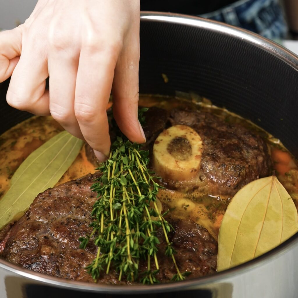 Adding bay leaves and thyme sprigs to the pot with beef shanks and braising liquid