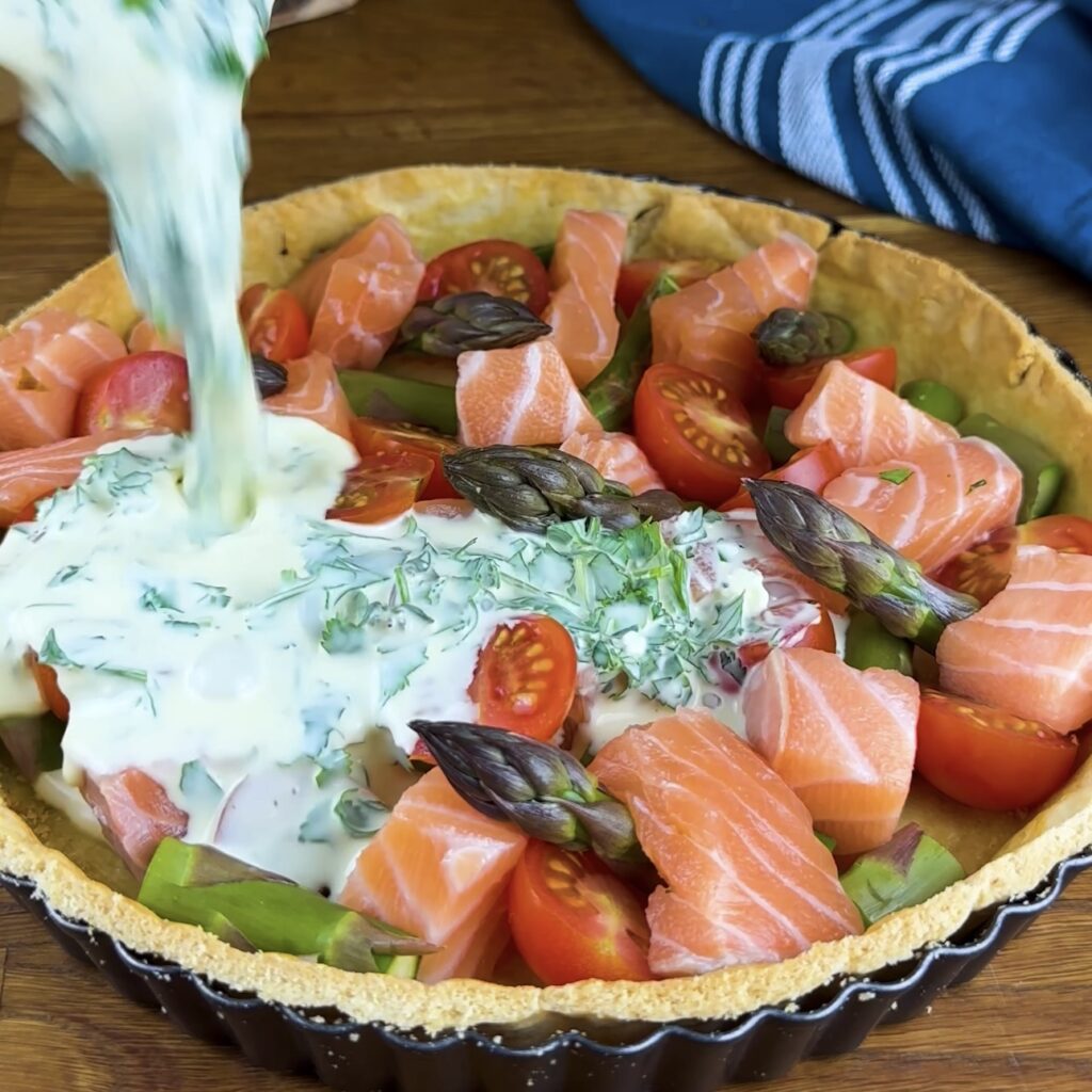 Pouring egg and cream mixture over salmon, asparagus, and tomatoes in tart base