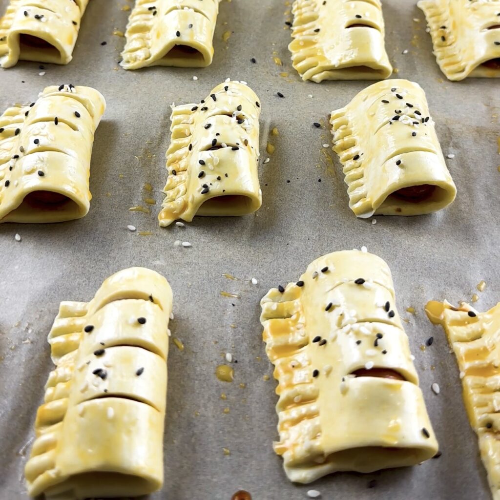 Mini sausage puffs on a baking tray brushed with egg wash and sesame seeds