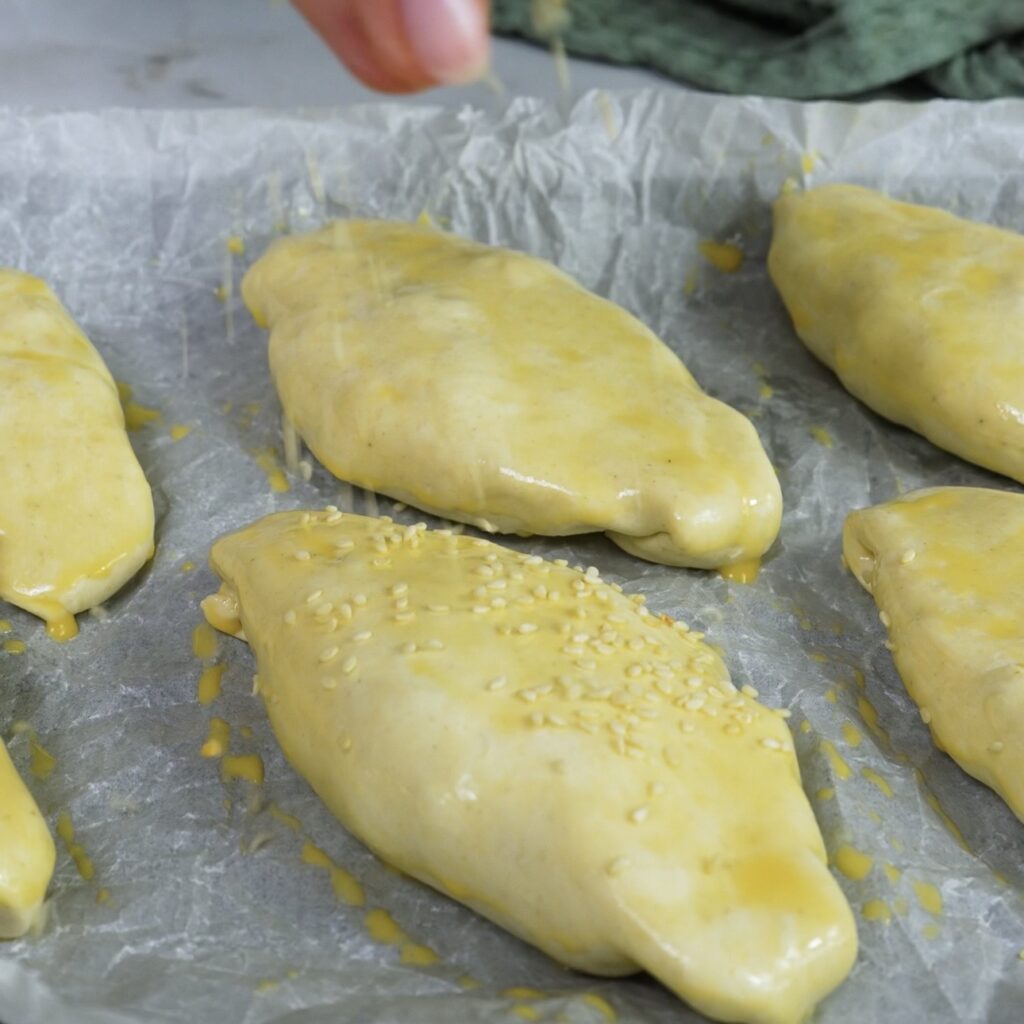 Puff pastry parcels seam side down on a baking tray, brushed with egg wash and sprinkled with sesame seeds.