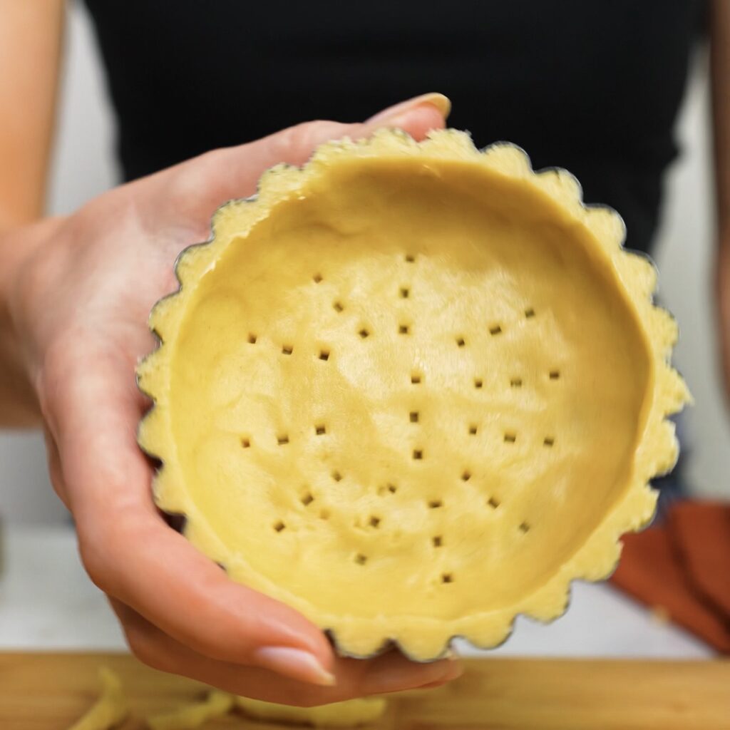 Hand holding a mini tart shell with shortcrust pastry pricked with a fork, ready for blind baking.