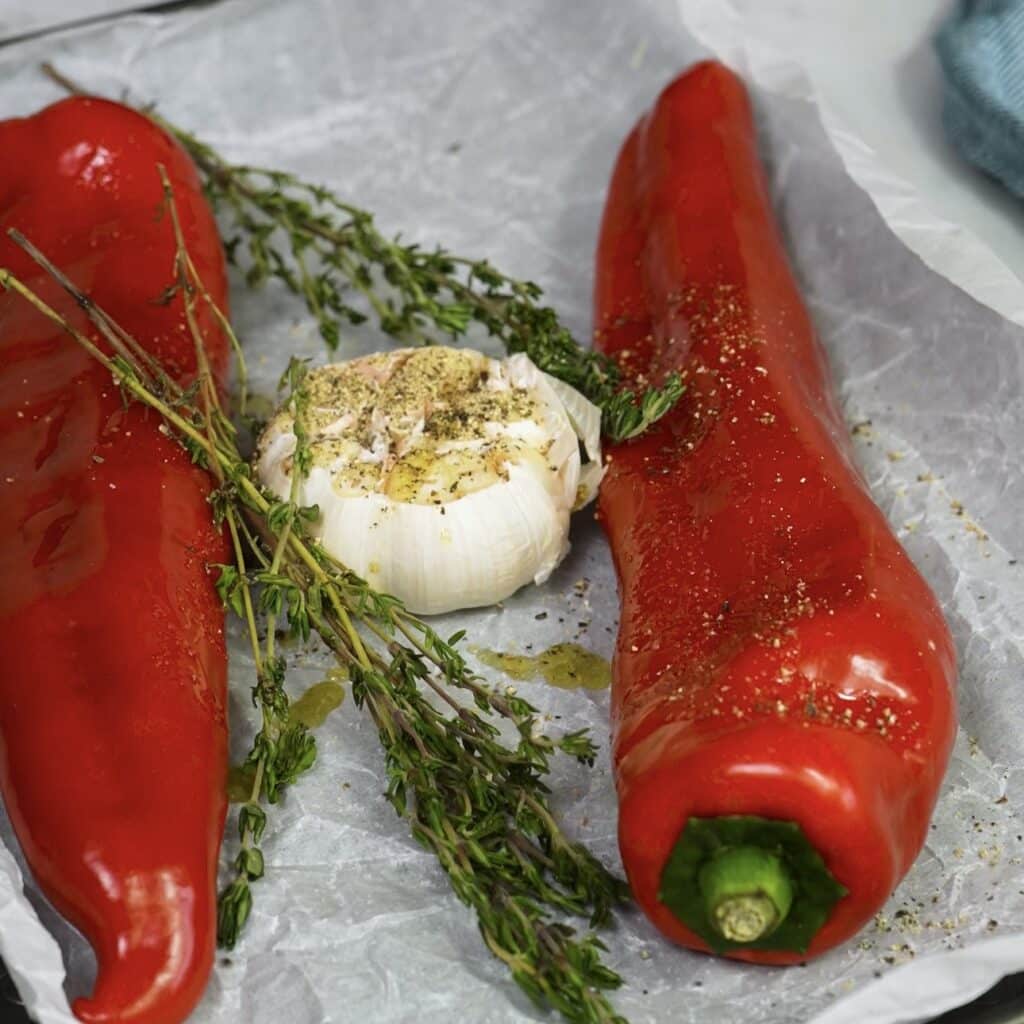 Red peppers, garlic and thyme on a baking tray ready for roasting for roasted pepper hummus