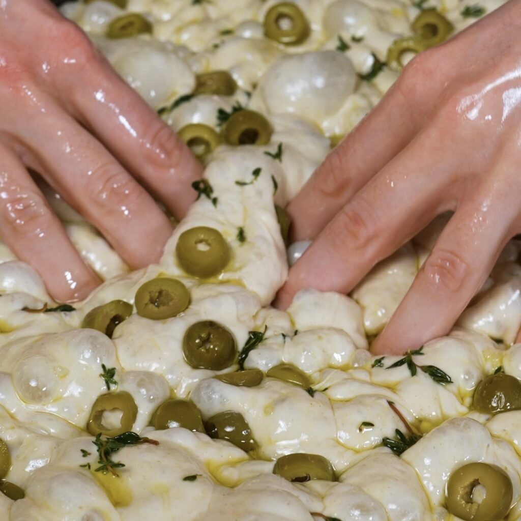 Dimpling focaccia dough with olives and thyme, pressing toppings into the surface