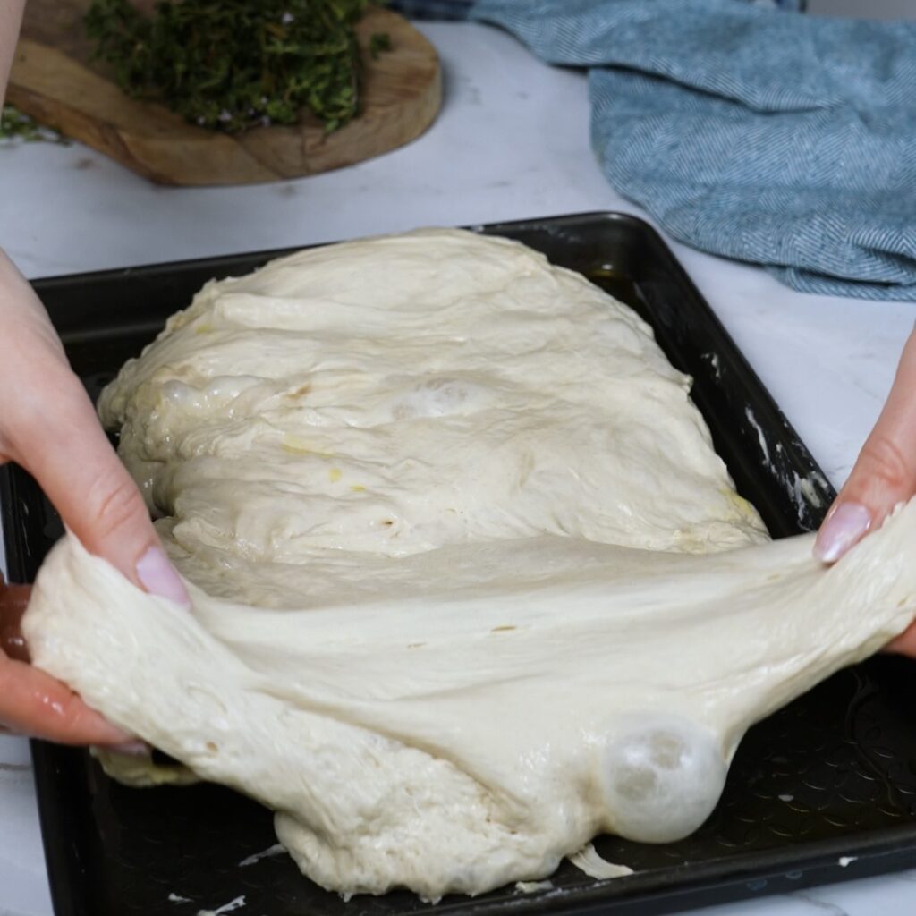 Stretching focaccia dough by hand in an oiled baking tray, gently spreading it to fill the pan evenly
