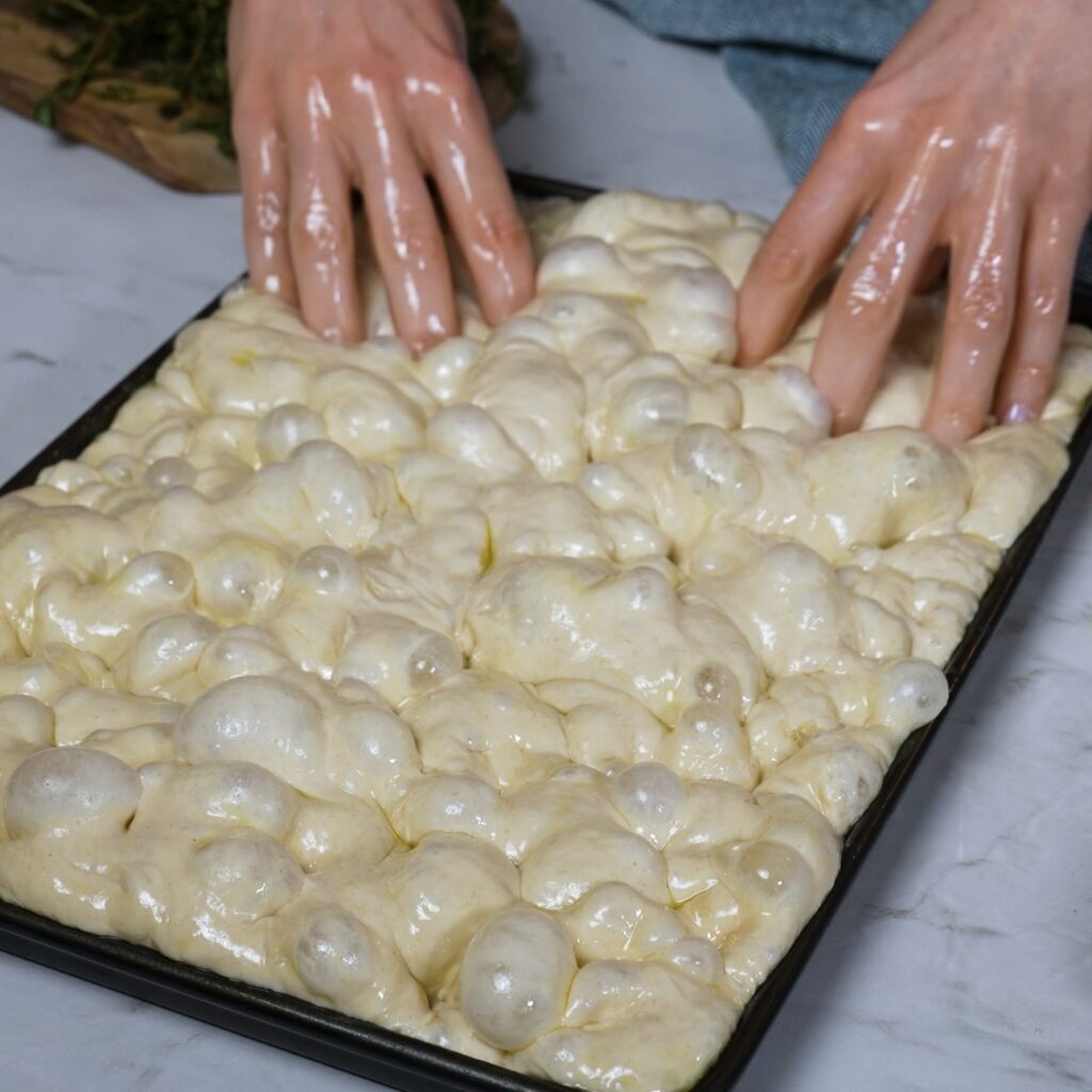 Dimpled focaccia dough in baking tray before adding toppings, with visible air bubbles and olive oil
