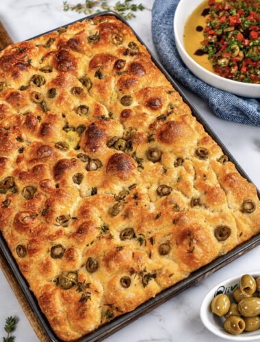Freshly baked focaccia in a baking tray with golden crust, olives, and thyme