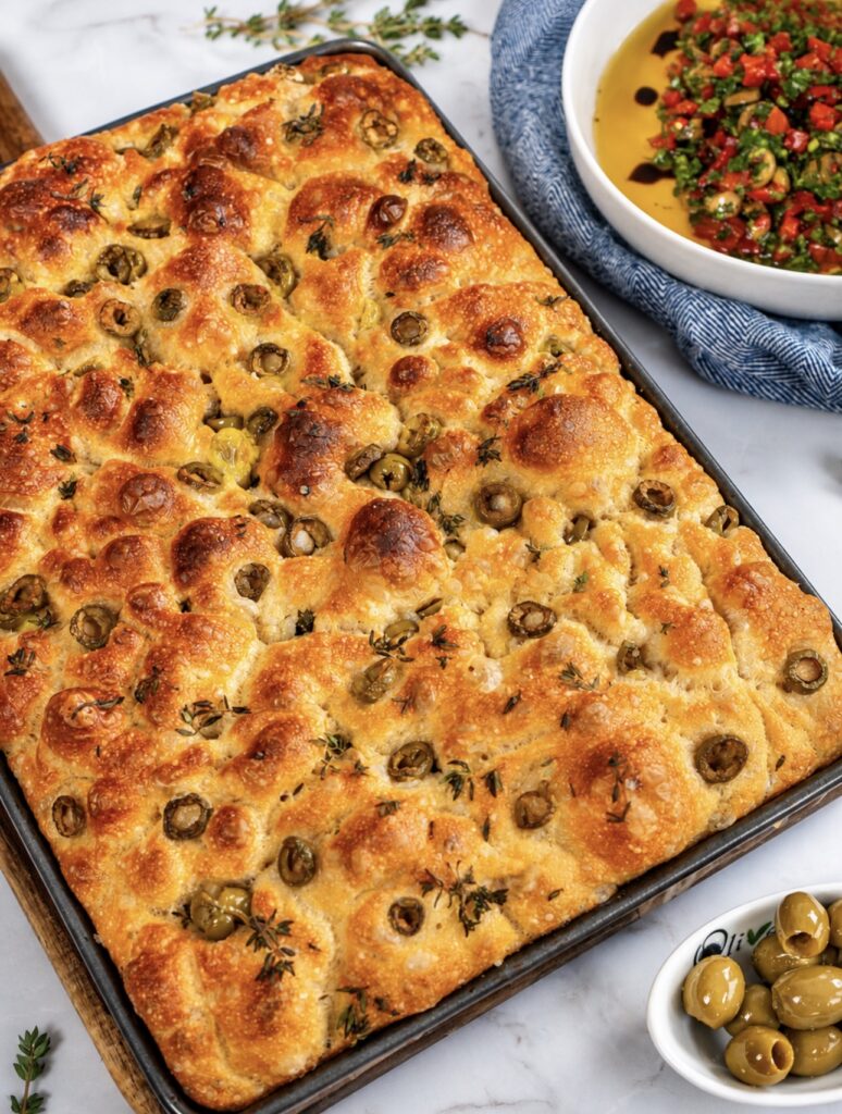 Freshly baked focaccia in a baking tray with golden crust, olives, and thyme