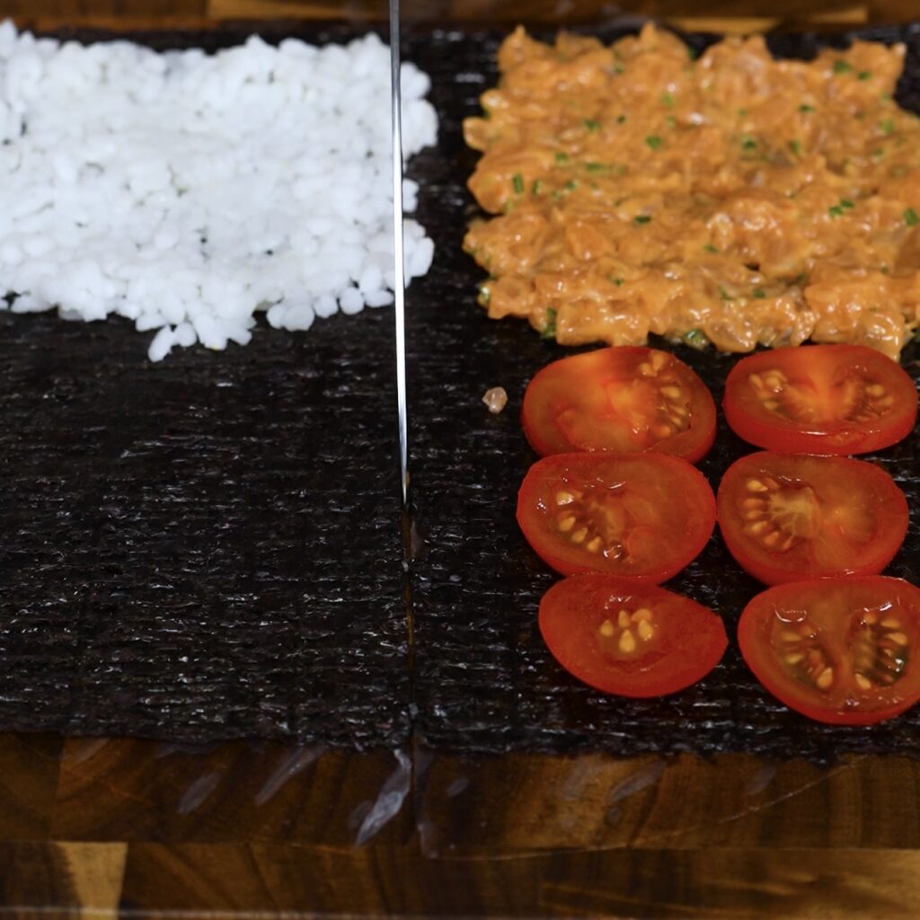 Sushi rice, salmon filling, and tomatoes layered on nori with a knife making the folding cut for sushi parcels