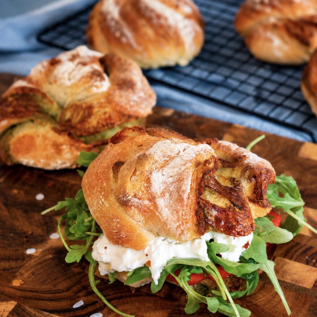Pesto, rocket, tomato, mozzarella, and burrata sandwich made with No-Knead Pesto Mozzarella Bread Twists on a wooden board.