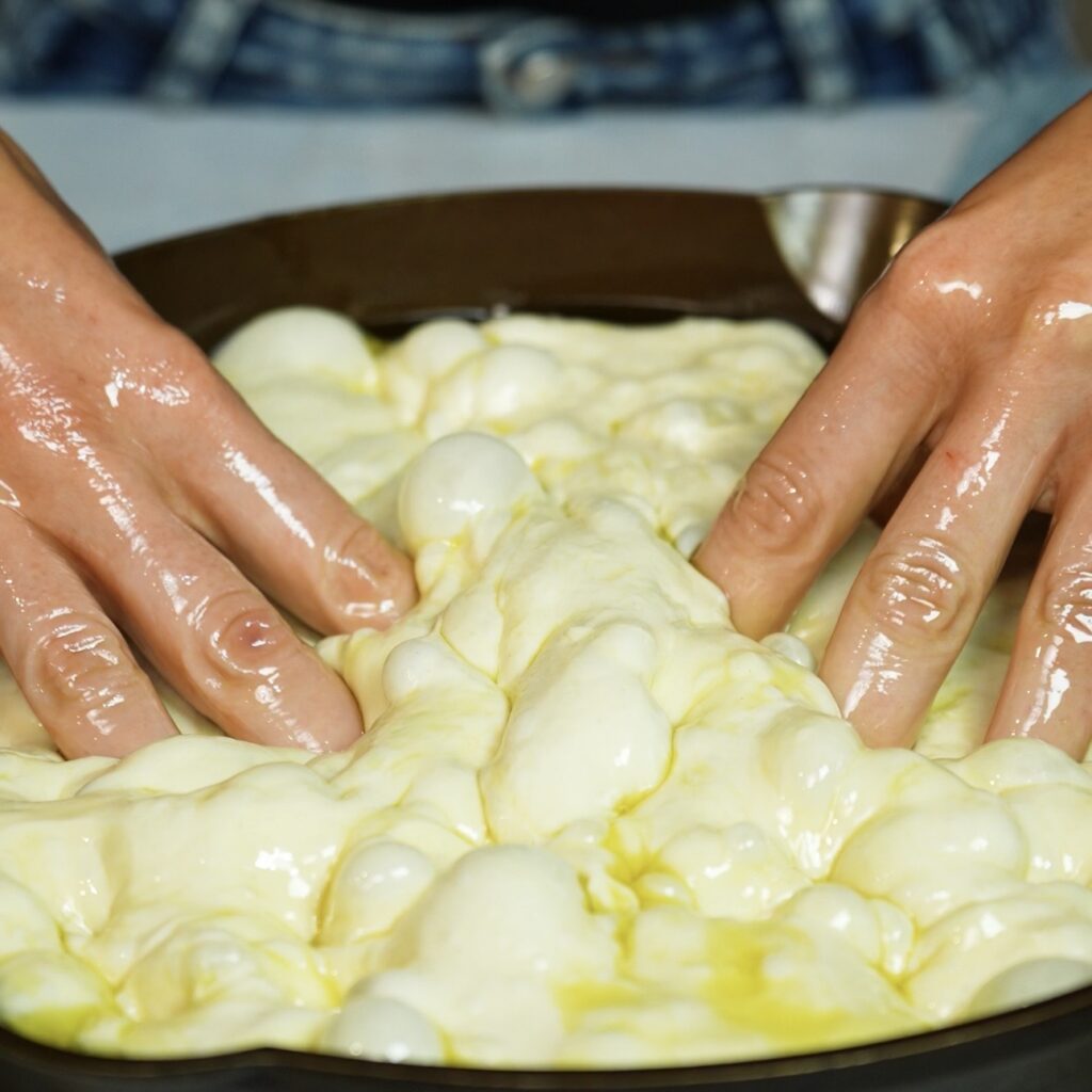 Hands dimpling focaccia dough in a round baking dish before adding pesto and cheese.