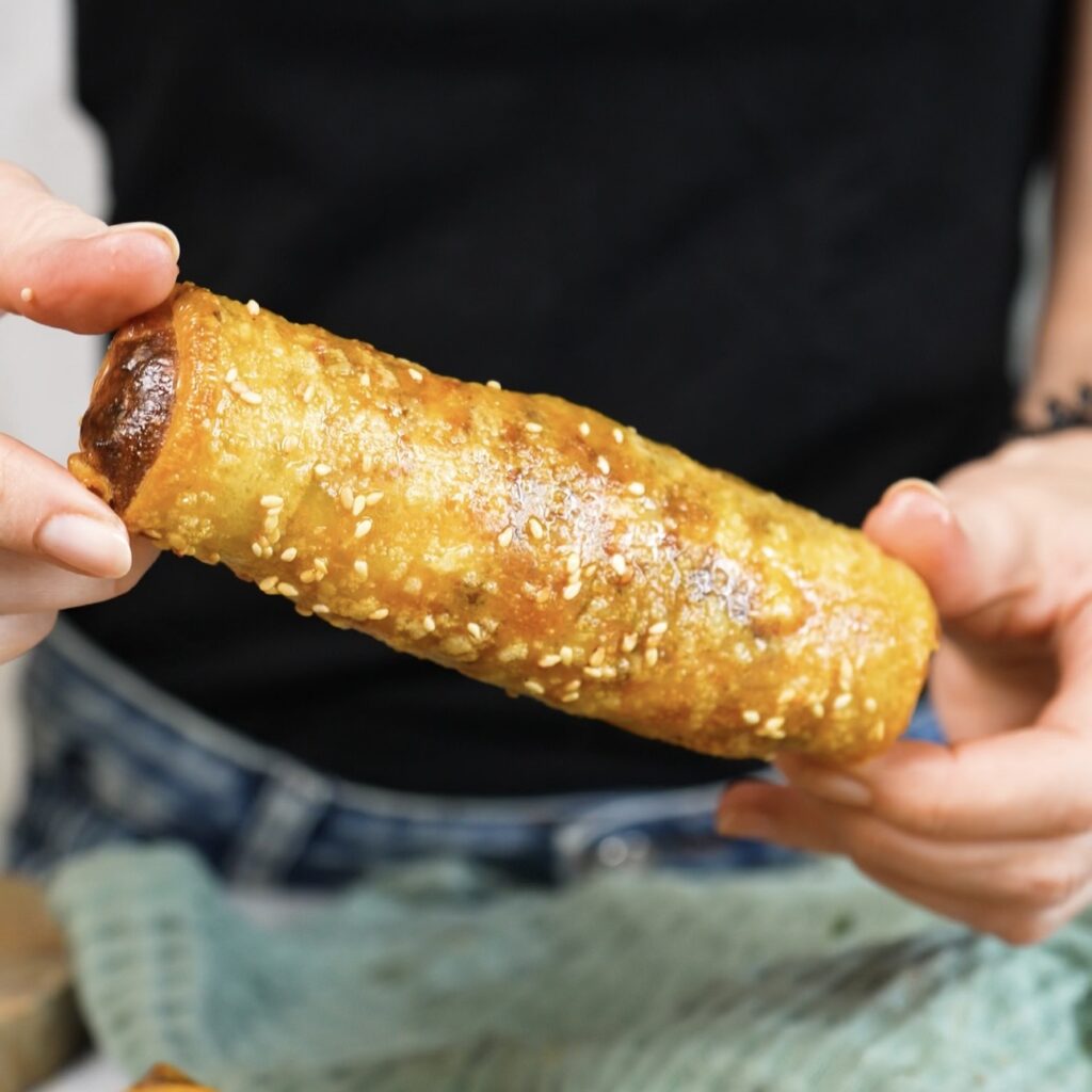 Hands holding a rice paper cheeseburger roll filled with beef, cheese, tomato and gherkins