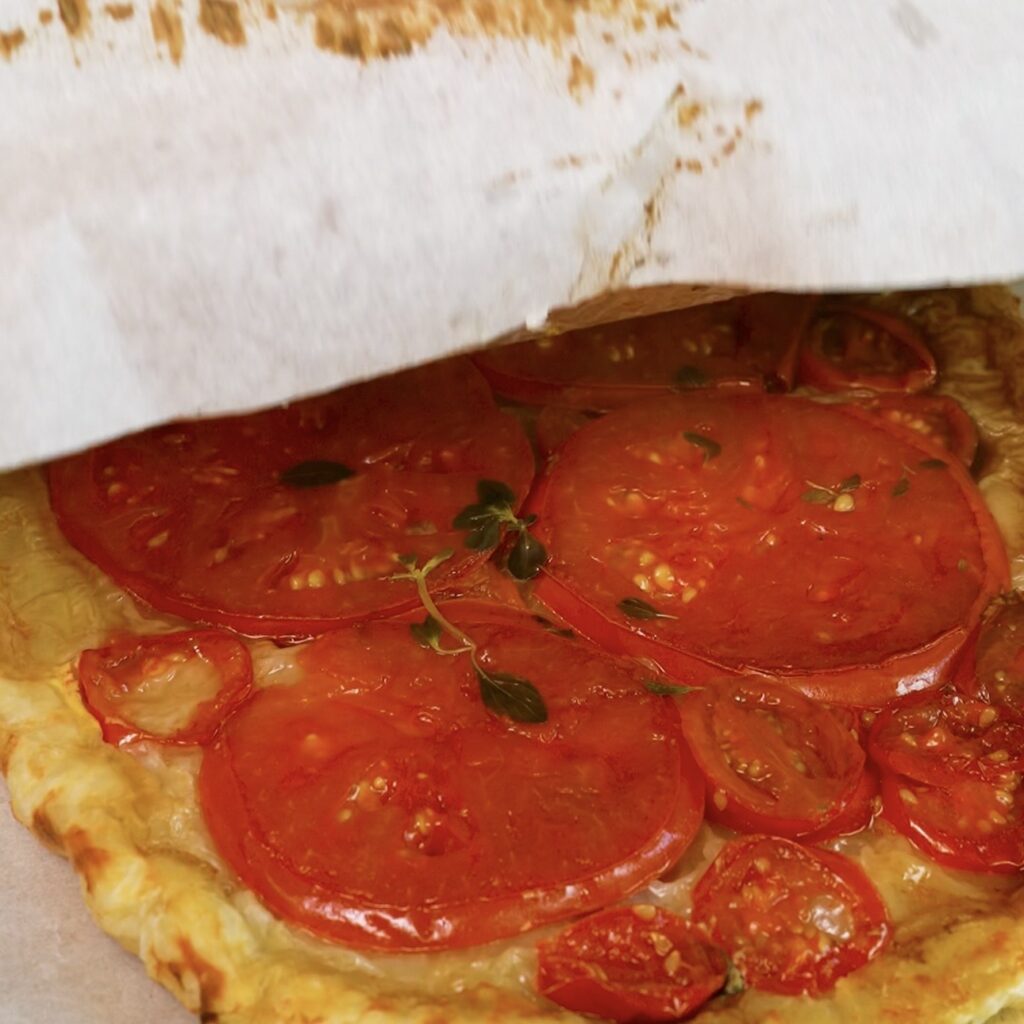 Hands peel parchment paper from a baked tomato and stracciatella upside-down tart.