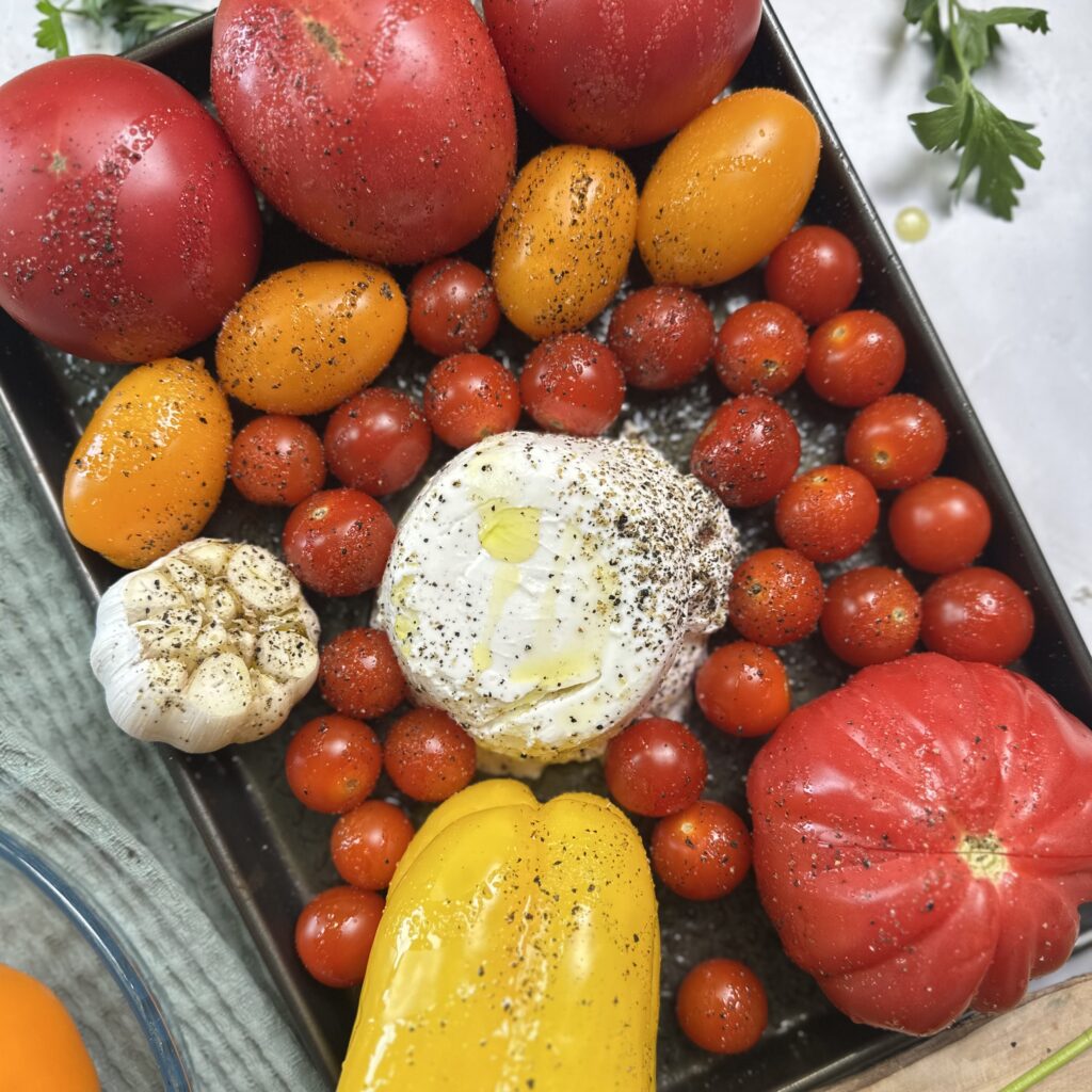 Tomatoes, cherry tomatoes, bell pepper, garlic bulb and ricotta arranged on a baking tray before roasting.