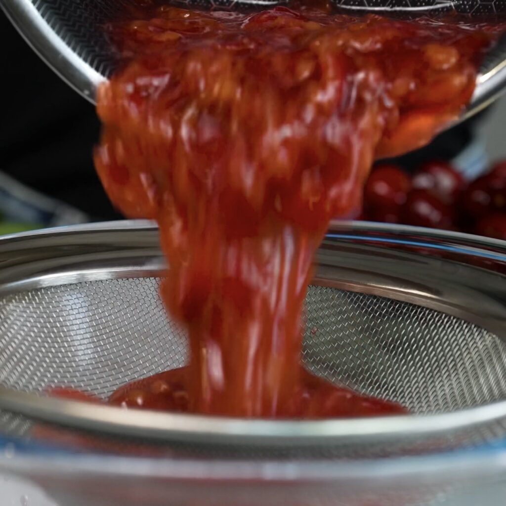 Cherry sauce being passed through a fine sieve into a bowl to make it smooth.