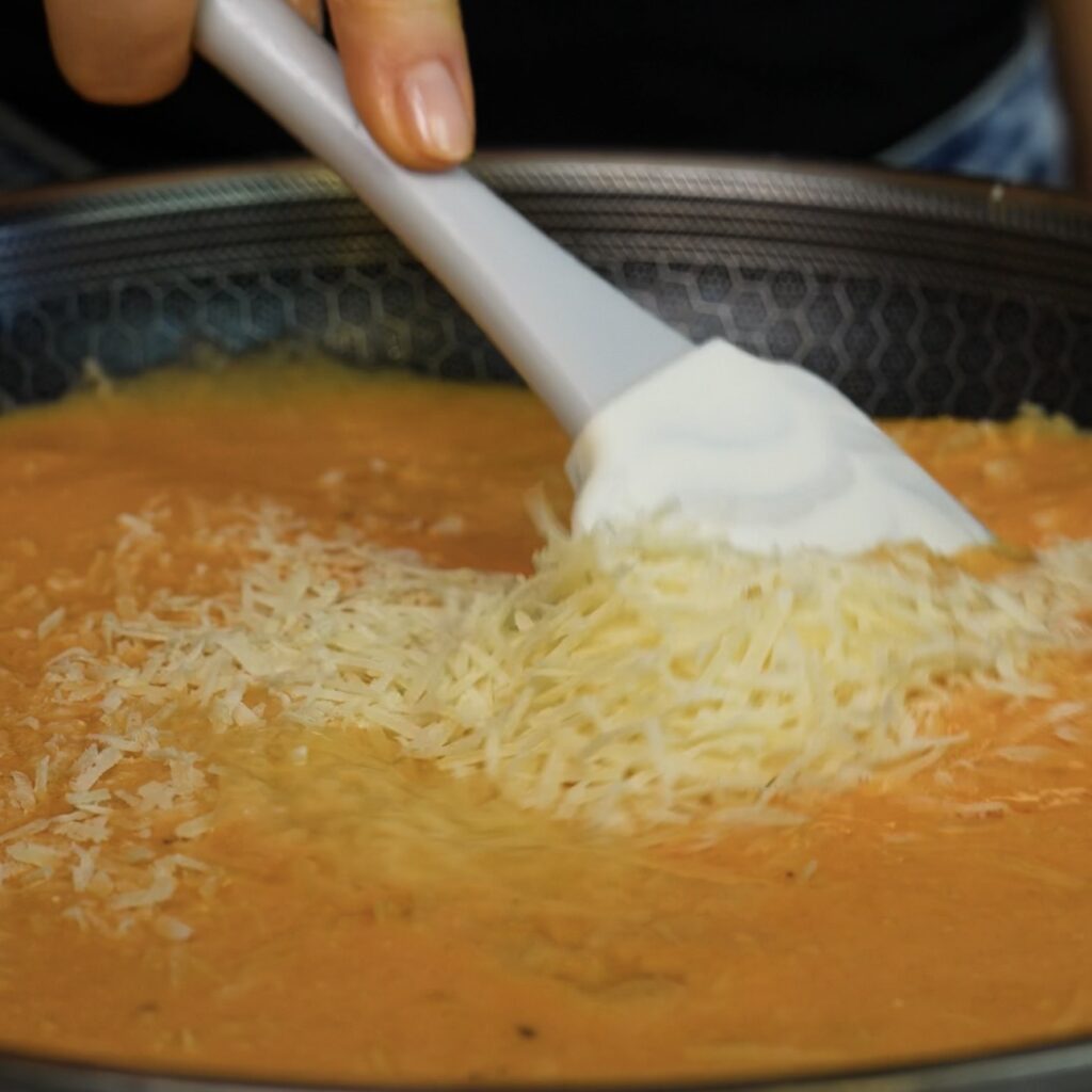 Grated Parmesan being stirred into the roasted tomato and ricotta sauce in a pan.