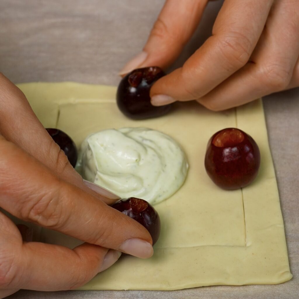 Pistachio yoghurt filling and pitted cherries arranged on a cut puff pastry square.