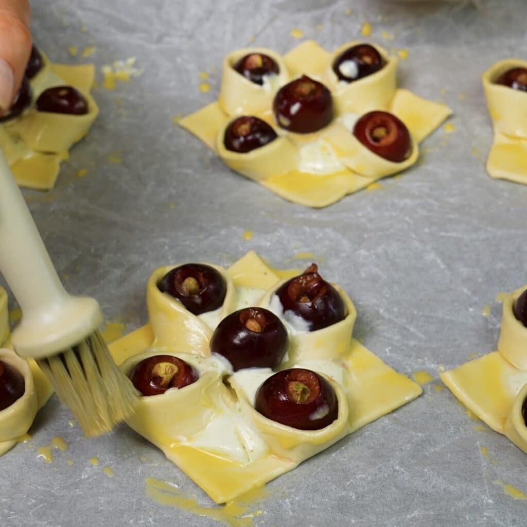 Shaped cherry and pistachio puff pastries being brushed with egg wash before baking.
