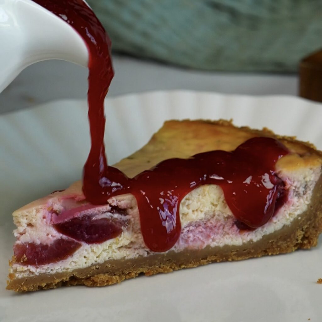 Pouring cherry sauce over a slice of Cherry Cheesecake