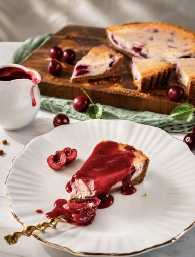 Slice of Cherry Cheesecake served on a plate with cherry sauce, with the remaining cheesecake in the background