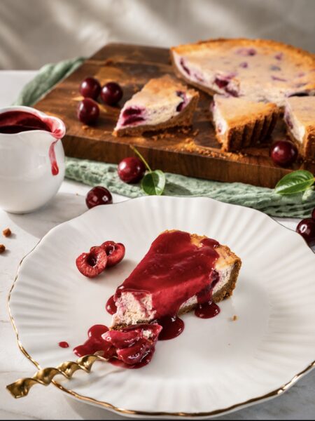 Slice of Cherry Cheesecake served on a plate with cherry sauce, with the remaining cheesecake in the background