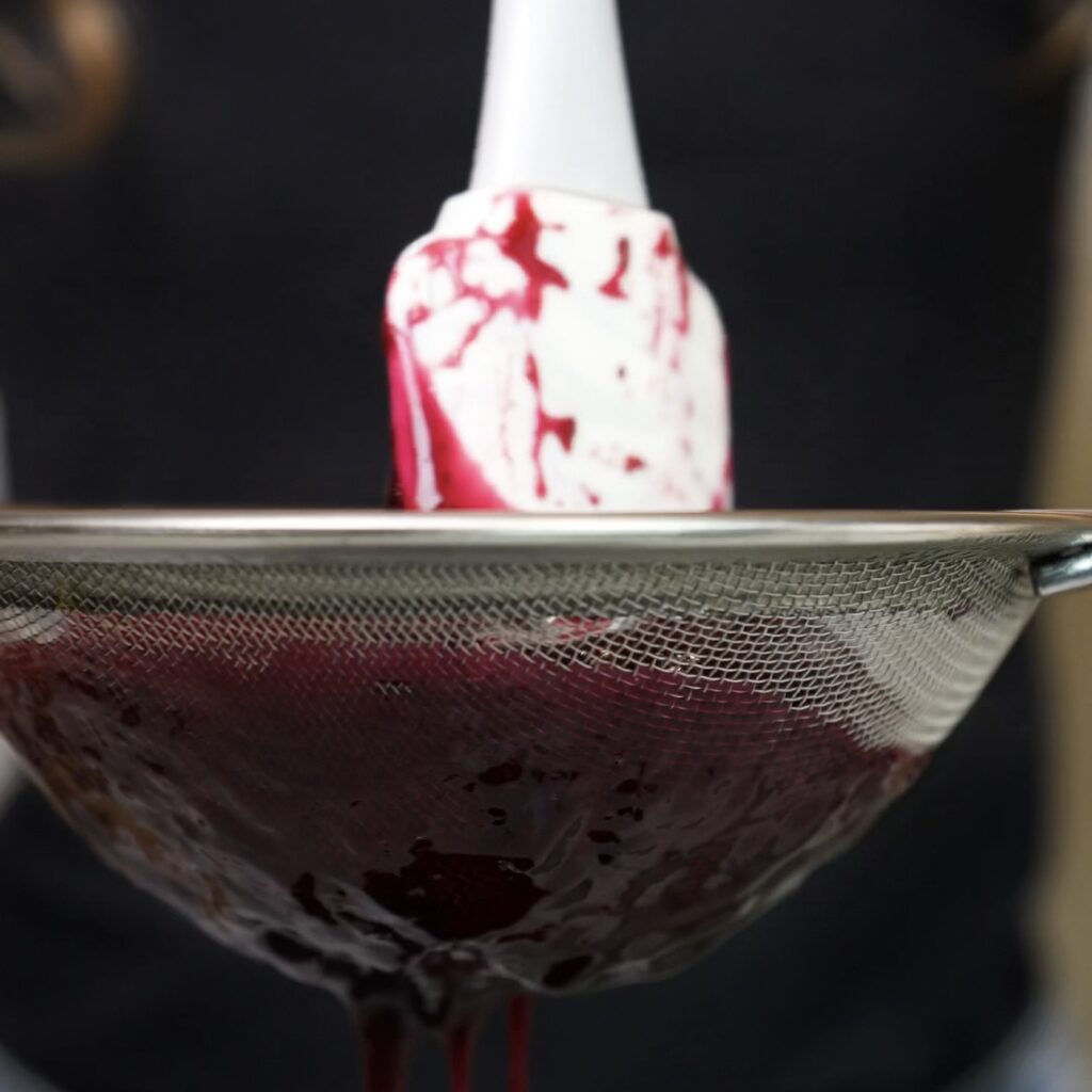 Blueberry sauce being passed through a fine-mesh sieve for a smooth consistency.
