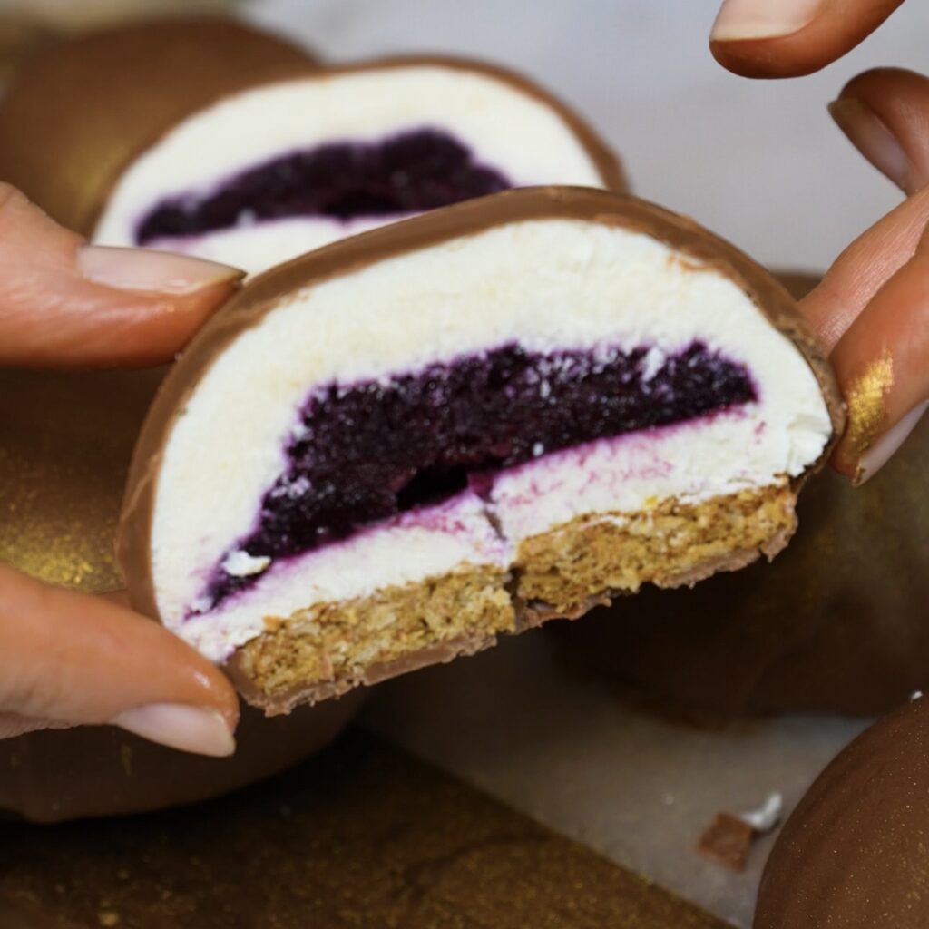 Hands holding half of a Blueberry Cottage Cheese Chocolate Sphere to reveal the creamy filling, blueberry centre and oat cookie base.