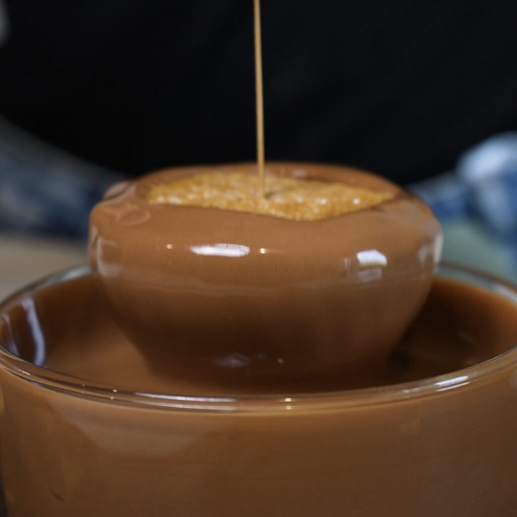 Frozen Blueberry Cottage Cheese Chocolate Sphere being dipped into melted milk chocolate while held by a toothpick.
